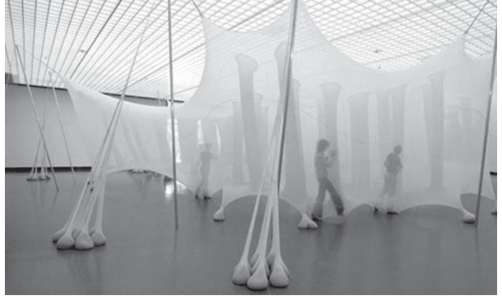 Fotografia em preto e branco de uma instalação artística composta por tecidos translúcidos esticados e suspensos formando tendas e estruturas tubulares flexíveis preenchidas na base, entre as quais pessoas caminham e interagem no interior de uma ampla galeria de arte.
