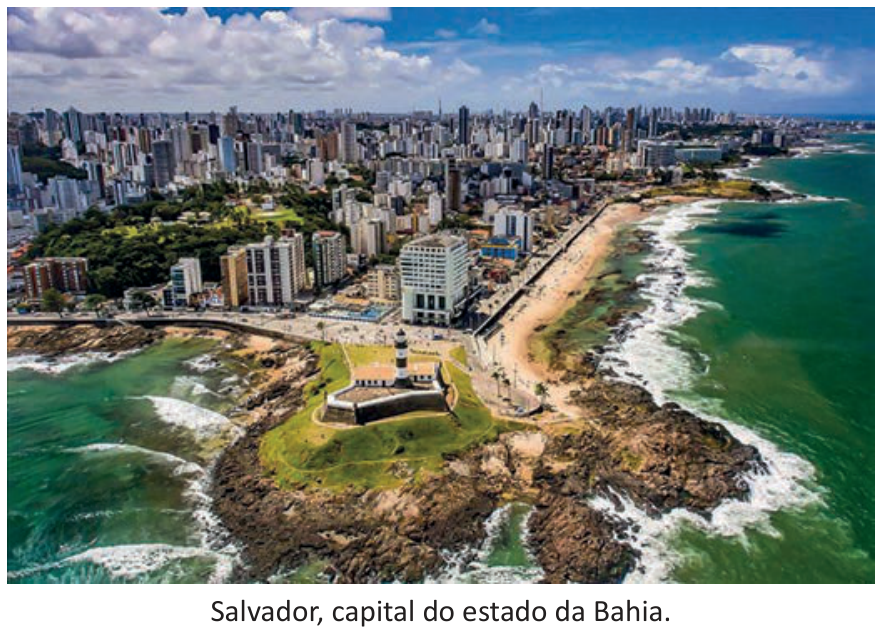 F1 Fotografia aérea mostrando o Farol da Barra em Salvador, Bahia, situado em uma ponta rochosa cercada pelo mar em primeiro plano. Ao fundo, estende-se a orla marítima e a malha urbana intensamente povoada por edifícios altos.