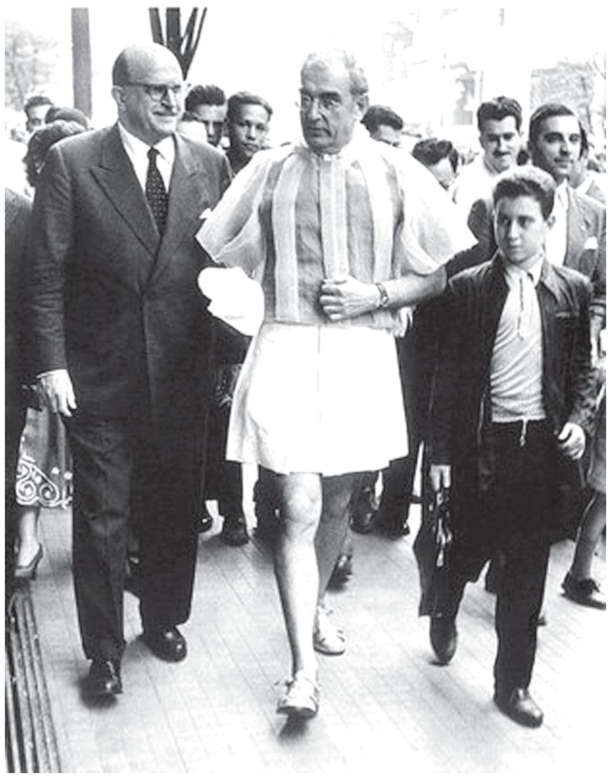 Fotografia em preto e branco do artista Flávio de Resende Carvalho caminhando no meio da calçada da Avenida Paulista com seu traje New Look, composto de saia curta plissada clara e blusa transparente de mangas bufantes, cercado por pedestres e homens vestidos com ternos escuros convencionais.