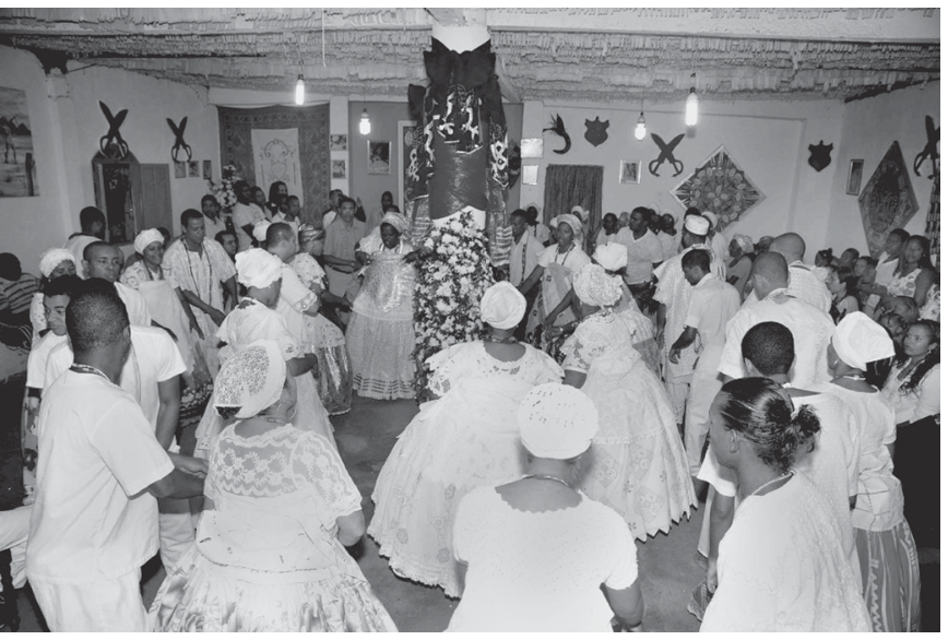 Fotografia em preto e branco retratando uma cerimônia em um terreiro de religião de matriz afro-brasileira, com praticantes vestidos com roupas brancas tradicionais, incluindo mulheres com saias rodadas e turbantes, reunidos e dançando ao redor de um pilar central decorado.
