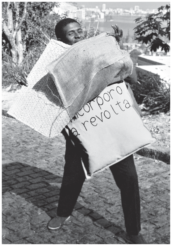 Fotografia em preto e branco de uma pessoa vestindo a obra "Parangolé", feita de tecidos e sacos, onde está escrito "incorporo a revolta". Ao fundo, observa-se uma paisagem ao ar livre com vista para a cidade.