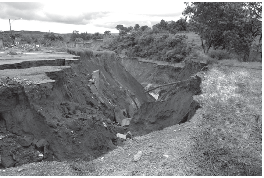 Fotografia em preto e branco de uma grande voçoroca que avança sobre uma área que parece pavimentada, exibindo uma enorme fenda de solo erodido com estruturas colapsadas e vegetação rasteira nas bordas