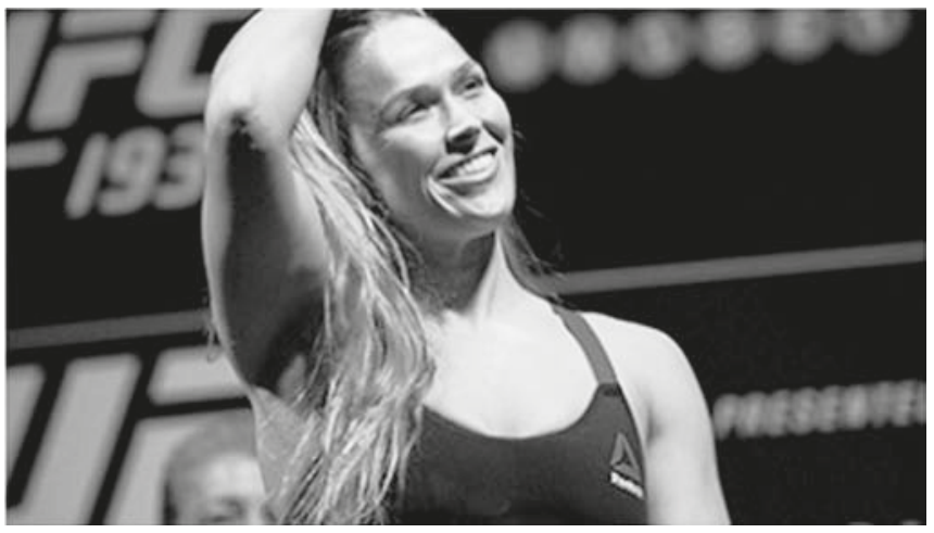 Fotografia em preto e branco da lutadora Ronda Rousey sorrindo e com a mão direita na cabeça, usando um top esportivo em frente a um painel do UFC.