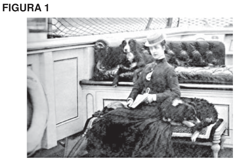 Fotografia em preto e branco da Princesa Alexandra sentada em um banco com cães, usando vestido longo e chapéu, segurando um livro aberto.