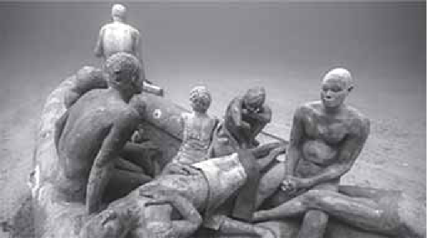 F1 Fotografia da escultura submarina "A balsa de Lampedusa", mostrando figuras humanas acinzentadas em tamanho real amontoadas sobre um bote no fundo do oceano.