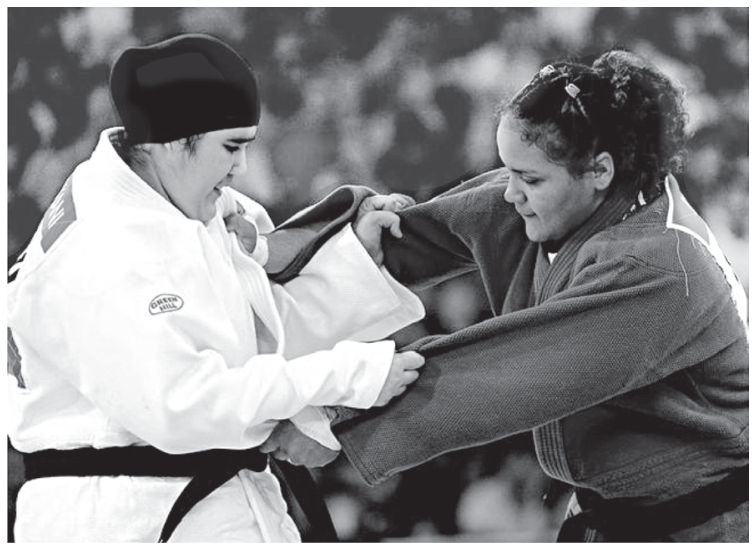 Duas atletas de judô disputando a luta; à esquerda, a atleta saudita veste quimono branco e usa uma touca escura justa cobrindo os cabelos (hijab adaptado); à direita, sua adversária veste quimono escuro. Ambas estão com as mãos segurando o quimono uma da outra.