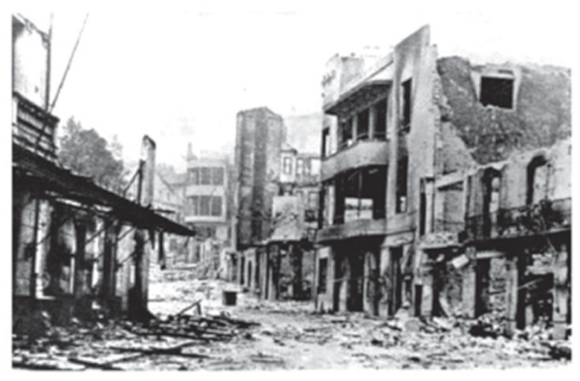 Fotografia em preto e branco de uma rua de Guernica em ruínas, com escombros espalhados e prédios destruídos após o bombardeio.