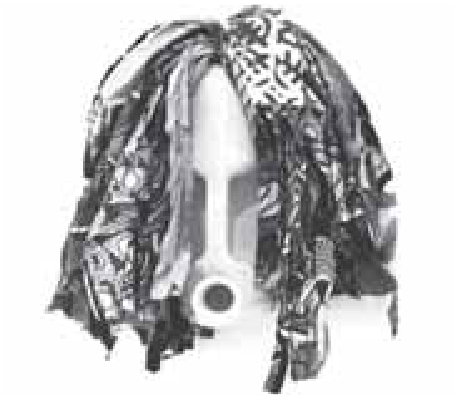 Escultura representando uma máscara com formato antropomórfico, composta por um recipiente plástico branco com orifício na parte inferior que lembra uma boca e fendas laterais, emoldurado por longas tiras de tecido estampado e escuro simulando cabelos.