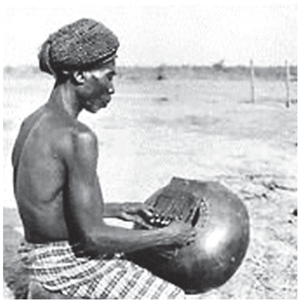 Fotografia em preto e branco de homem da cultura lupa visto de perfil, sem camisa e com turbante, tocando uma kalimba ou lamelofone sobre o colo em campo aberto.