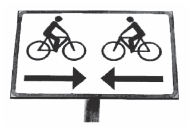 Placa retangular de sinalização de ciclistas. Na parte superior, há dois ciclistas sobre bicicletas virados um em direção ao outro; na parte inferior, abaixo de cada ciclista, há uma seta horizontal apontando para o centro da placa, ou seja, a seta da esquerda aponta para a direita e a da direita aponta para a esquerda.