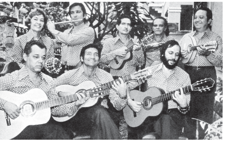 Fotografia em preto e branco de um grupo de choro com oito integrantes vestidos com camisas de estampa idêntica. Três músicos estão sentados na frente tocando violão; atrás deles estão cinco músicos em pé segurando ou tocando, da esquerda para a direita: um afoxé, uma flauta transversal, um cavaquinho, um pandeiro e um bandolim.