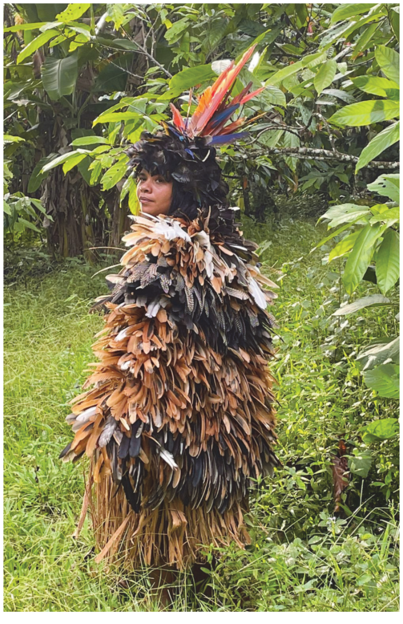 Fotografia de Glicéria Tupinambá vestindo a obra Assojaba Tupinambá, um manto tradicional confeccionado inteiramente com penas, em meio a uma área de vegetação.