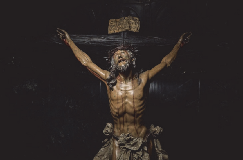 Detalhe de uma escultura em madeira de Cristo crucificado, com os braços estendidos e presos à cruz, coroa de espinhos, cabeça inclinada para cima, boca entreaberta, torso nu com detalhes anatômicos e tecido amarrado à cintura. Acima da cabeça, há uma placa com inscrições.