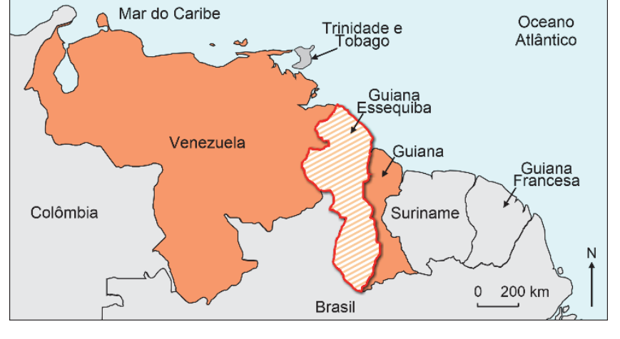 F1 Mapa da porção norte da América do Sul evidenciando a Venezuela e a Guiana, com destaque em linha vermelha e hachuras para a região disputada da Guiana Essequiba. Ao redor, figuram a Colômbia, o Brasil, o Suriname, a Guiana Francesa, Trinidad e Tobago, além do Mar do Caribe e do Oceano Atlântico, com barra de escala gráfica e rosa dos ventos indicando o Norte.
