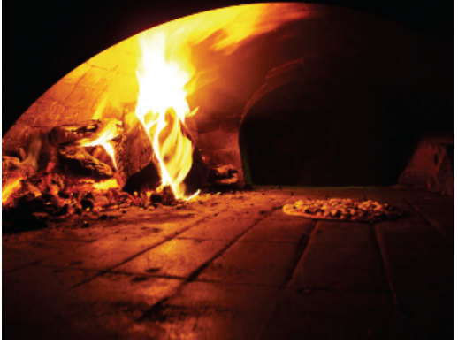 F1 Fotografia do interior de um forno a lenha com paredes escuras, lenha em chamas à esquerda e uma pizza à direita.