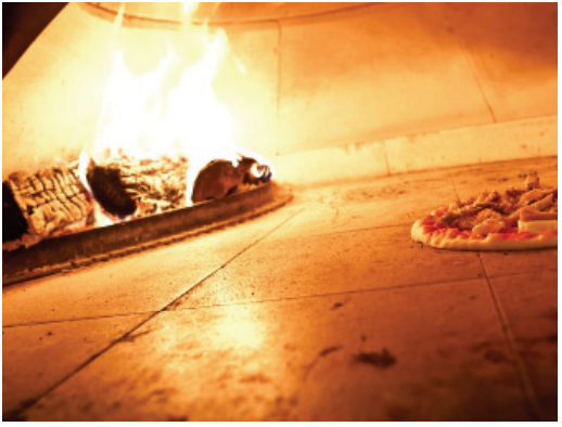 F2 Fotografia do interior de um forno a lenha com paredes claras, lenha em chamas à esquerda e uma pizza à direita.