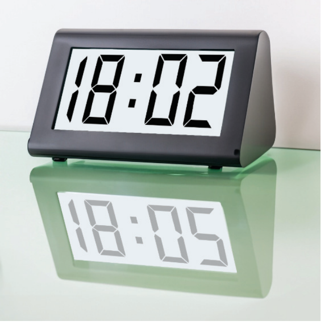 Fotografia de um relógio digital sobre uma mesa de vidro. O visor marca 18:02, e seu reflexo na superfície do vidro apresenta os algarismos correspondentes a 18:05.