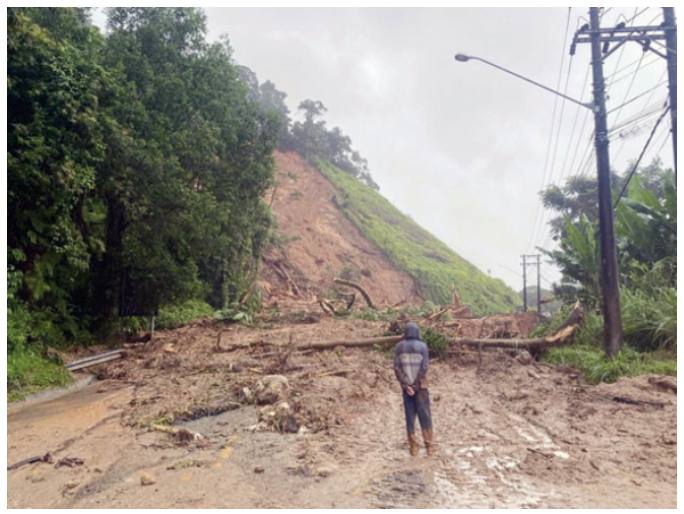 Fotografia de uma estrada totalmente obstruída por lama, detritos e troncos de árvores decorrentes de um deslizamento de terra na encosta íngreme ao fundo; uma pessoa com capa de chuva observa a destruição de costas.