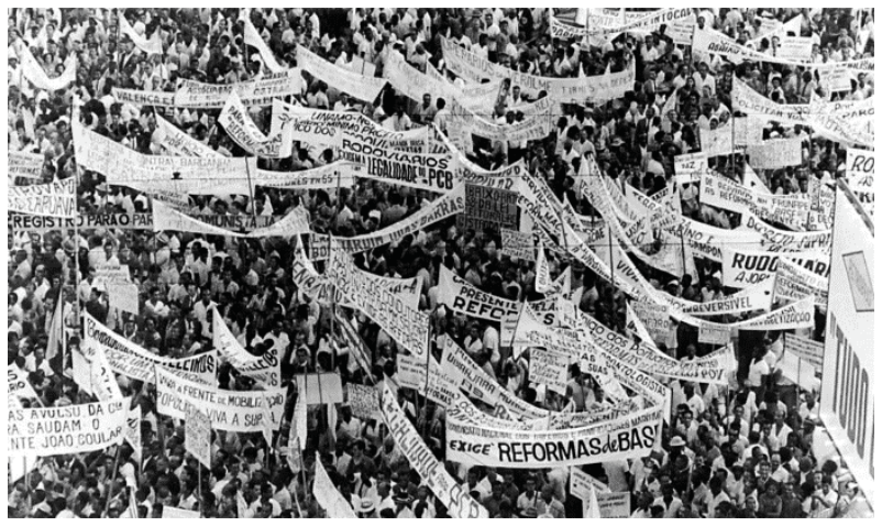 Fotografia aérea mostrando uma grande multidão portando diversas faixas e cartazes com reivindicações políticas e trabalhistas.