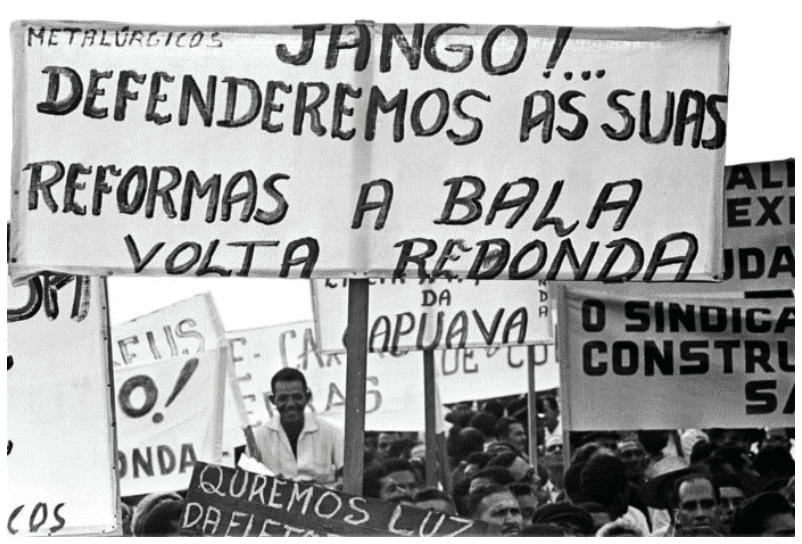 Fotografia em meio à multidão destacando um cartaz sustentado em uma haste de madeira com a frase: "METALÚRGICOS JANGO!... DEFENDEREMOS AS SUAS REFORMAS A BALA VOLTA REDONDA". Ao fundo, outras faixas e pessoas presentes no comício.