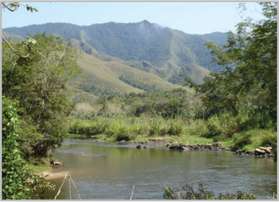 Fotografia de um rio largo de águas calmas em primeiro plano, ladeado por vegetação ciliar, com morros e montanhas íngremes ao fundo.