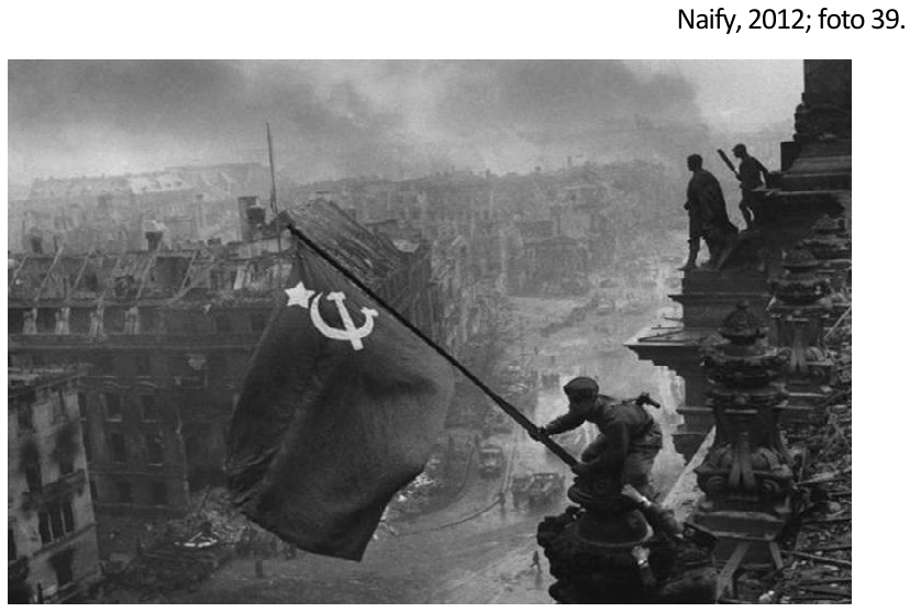 Fotografia em preto e branco de um soldado erguendo uma bandeira soviética com estrela, foice e martelo no alto de um edifício, com outros soldados à direita e edifícios destruídos de Berlim ao fundo.