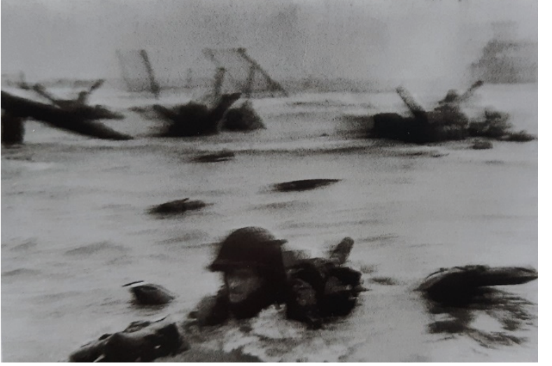 Fotografia em preto e branco de um soldado de capacete parcialmente submerso na água, com obstáculos de desembarque ao fundo, na praia de Omaha.