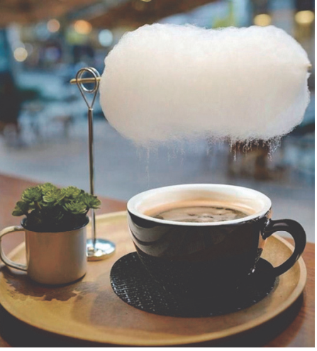 Fotografia de uma xícara de café sobre um pires, em uma bandeja de madeira. Acima da xícara, um suporte mantém um algodão-doce branco suspenso, com aparência de nuvem. Ao lado da xícara há um pequeno vaso com uma planta.